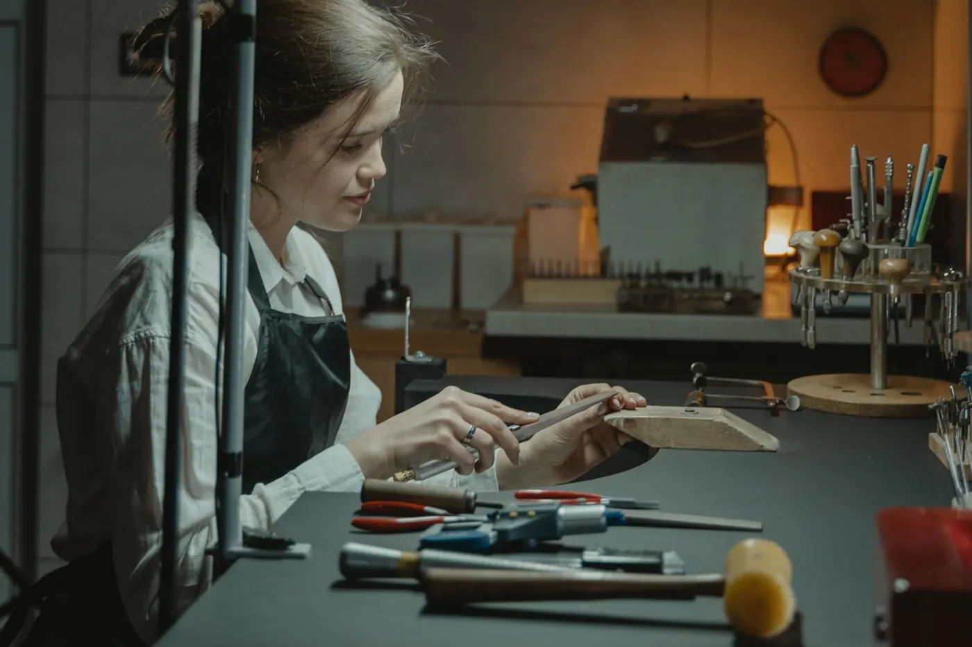 A goldsmith at a jeweller&rsquo;s bench under a work lamp, filing a ring, with hammers and pliers laid out in front of her