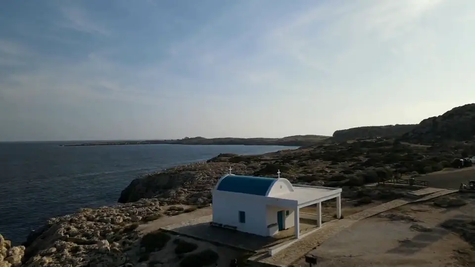 Still from an aerial film: a white chapel with a blue dome on a rocky cape above the sea