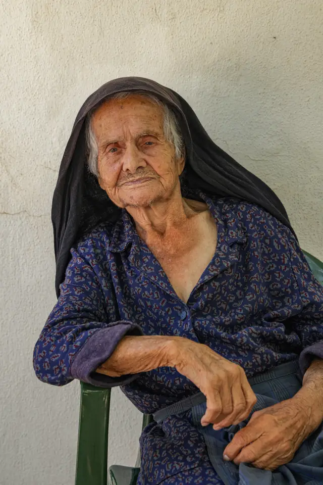 Elderly woman in a black headscarf and blue dress seated against a whitewashed wall
