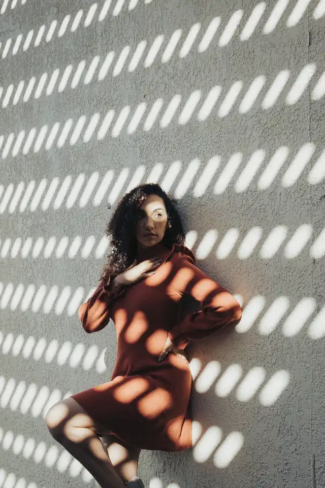 Woman in a rust-coloured dress leaning on a white wall striped with sunlight through a shutter