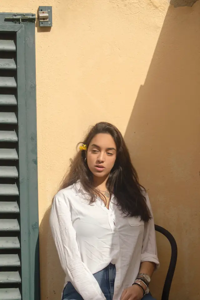 Young woman in a white shirt sitting against a sunlit yellow wall beside green shutters